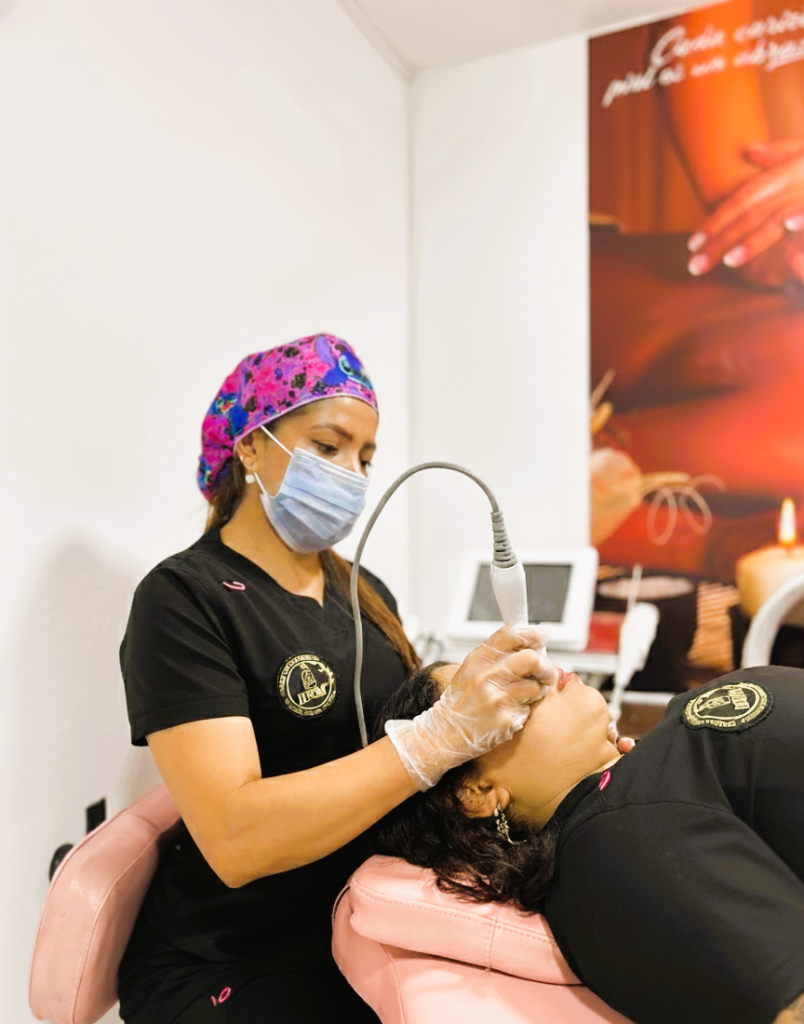 Profesional de estética realiza tratamiento facial avanzado a paciente en camilla rosa, con enfoque nítido en el equipo y el procedimiento.